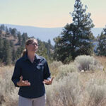 A woman wearing glasses and a dark shirt stands in a dry, grassy area