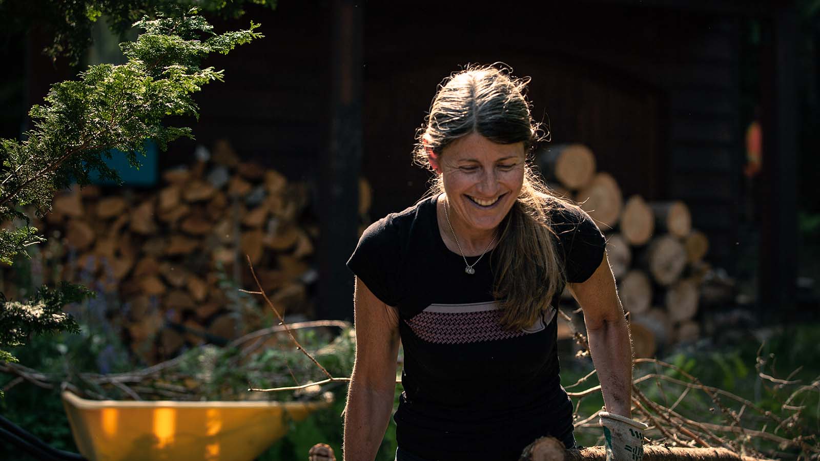 A woman smiling while working outdoors, with a pile of firewood in the background.