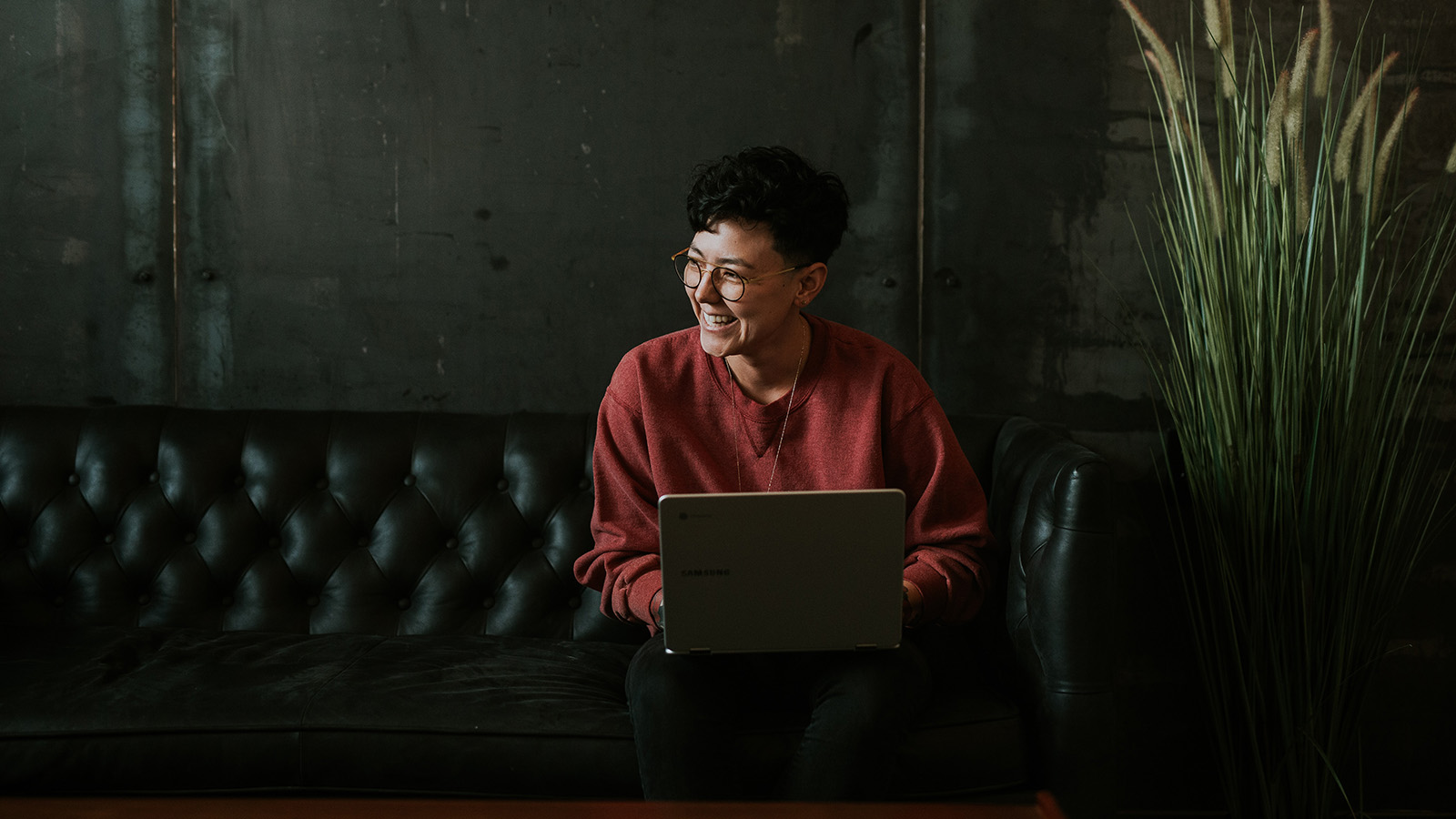 A person with glasses and a red sweatshirt is smiling while using a laptop