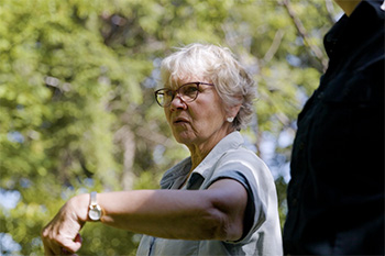 An elderly woman with glasses stands outdoors