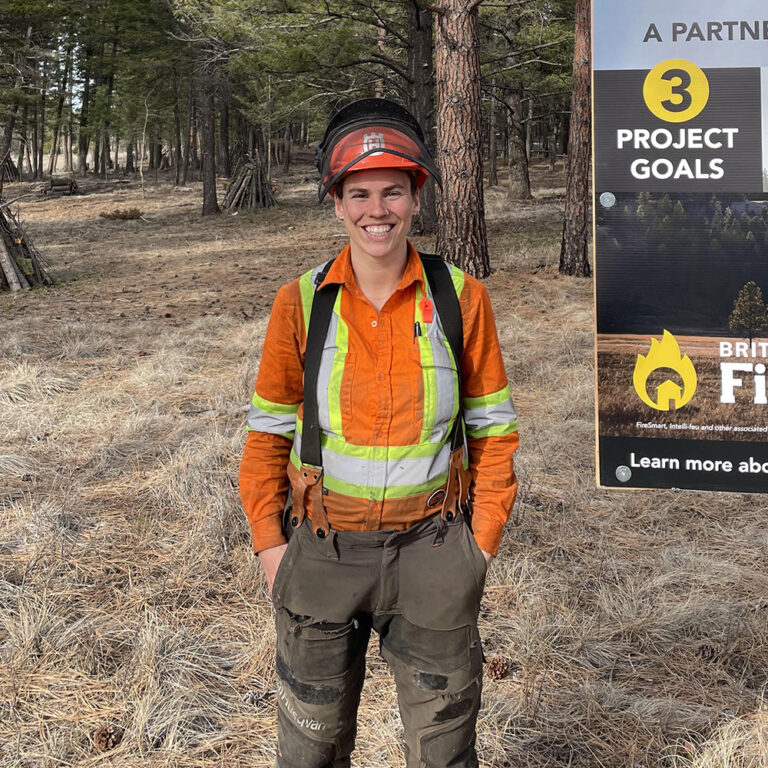 A person wearing safety gear stands in a forested area, smiling at the camera.