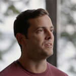 A man with short hair looks off to the side, with a blurred outdoor background.