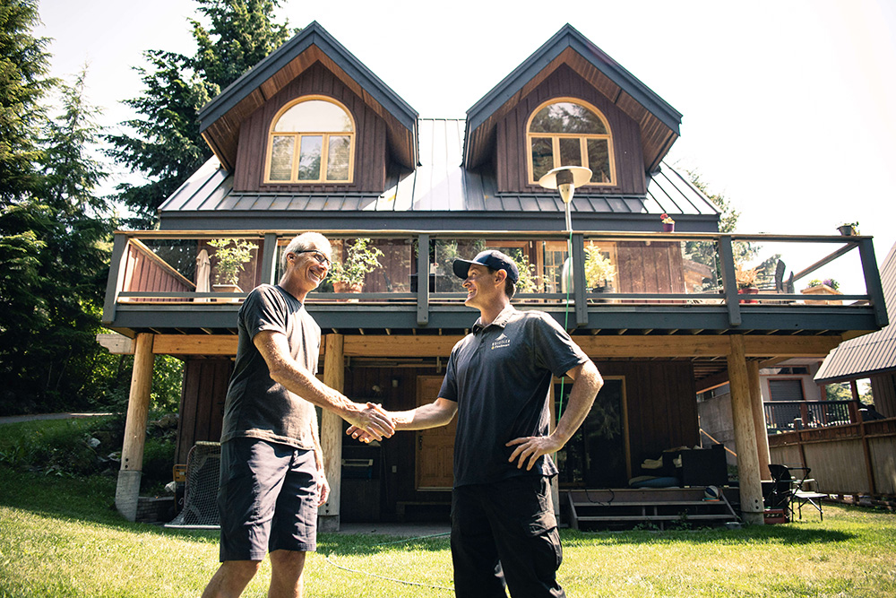 Two men are shaking hands in front of a two-story house with a large deck.