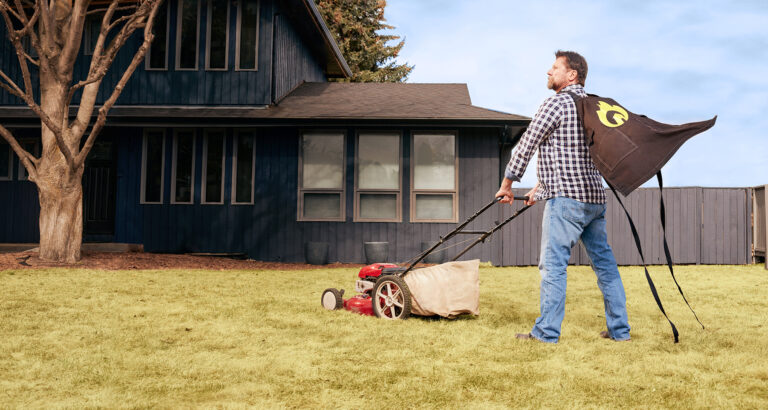 A person wearing a cape is mowing the lawn in front of a house.