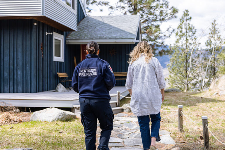 Two women, one wearing a "Penticton Fire Rescue" jacket, walk towards a house.