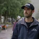 A man in a black cap and jacket stands outdoors.
